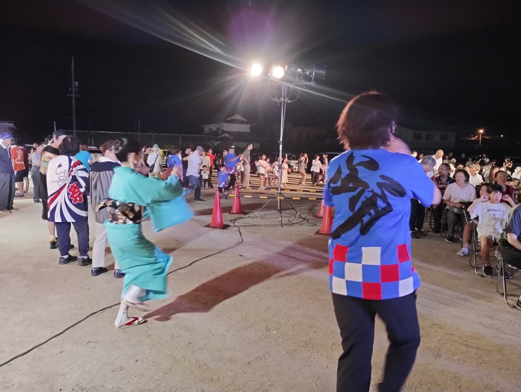 夜の運動場で、そろいのTシャツや浴衣の人たちが輪になって踊っている