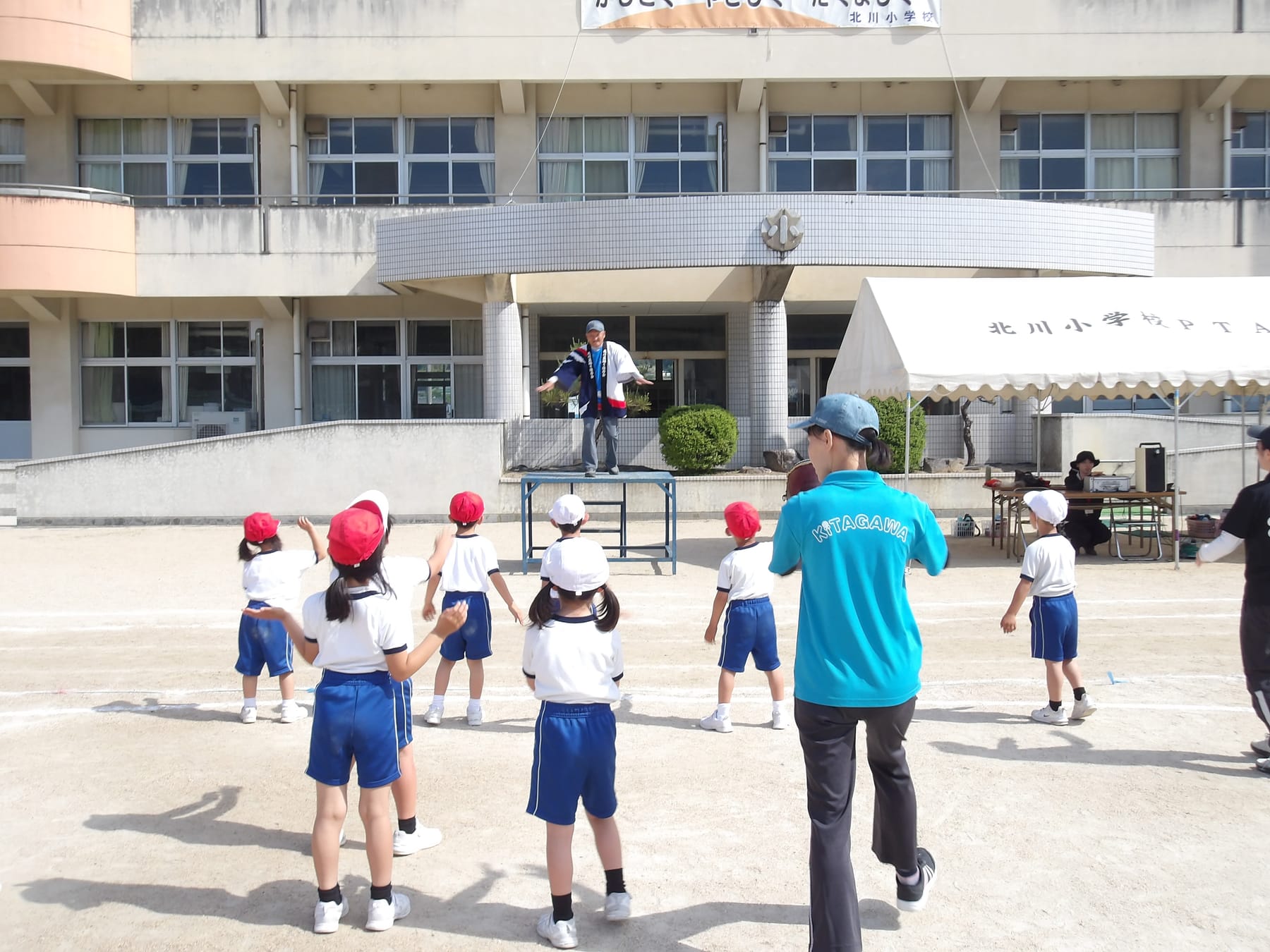 校舎の前の台に立つ保存会の会員にあわせて、子どもたちが踊っている（後ろ姿）
